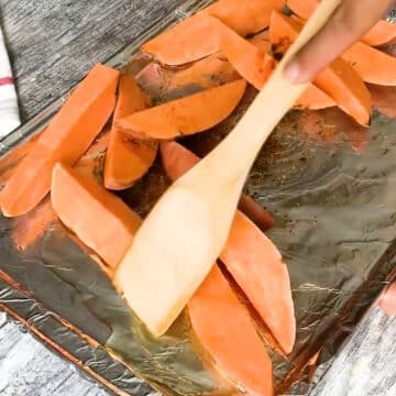 Sweet potatoes being tossed in oil and seasoning with a wooden spoon.