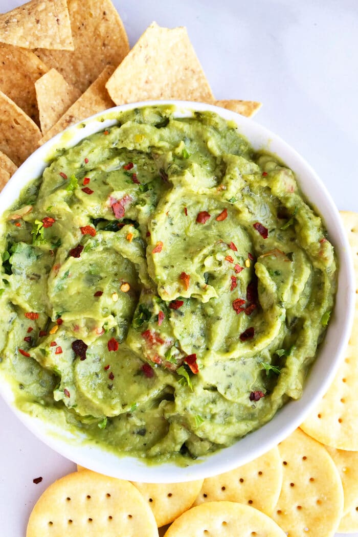 Spicy Mexican avocado dip in white bowl on large dish with crackers and tortillas. 