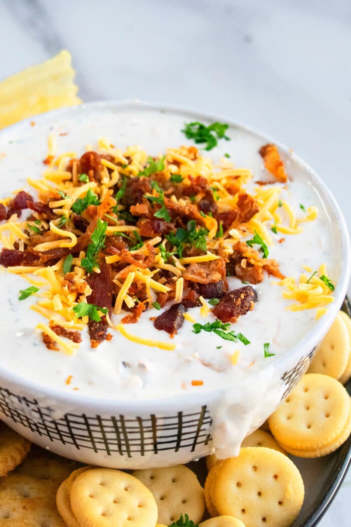 Bowl of creamy crack dip with bacon and cheddar topping on dish with crackers. 