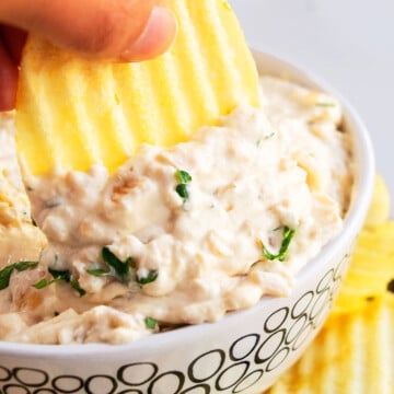 Potato Chip Being Dipped Into Best Easy Caramelized French Onion Dip in White Bowl.
