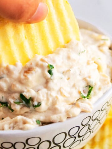 Potato Chip Being Dipped Into Best Easy Caramelized French Onion Dip in White Bowl.