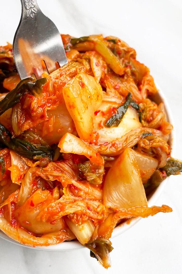 Closeup Shot of Fork Packed With Red Fermented Pickled Korean Cabbage in White Bowl on White Background.