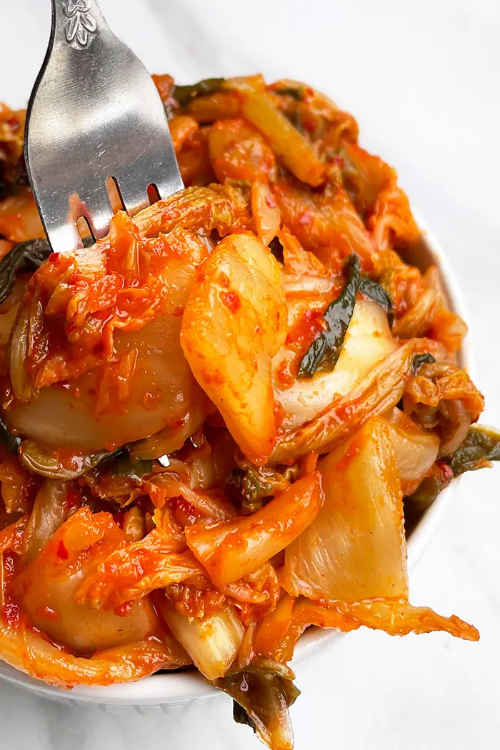 Closeup Shot of Fork Packed With Red Fermented Pickled Korean Cabbage in White Bowl on White Background