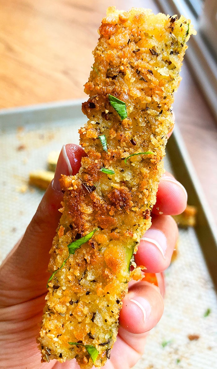 Holding a Breaded Zucchini Stick in Hand Over a Baking Tray