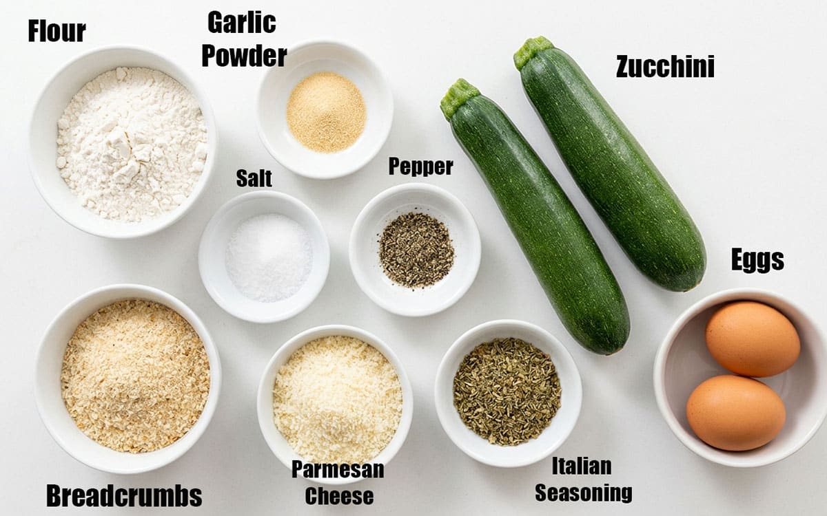 Ingredients for recipe on white background- zucchini, seasonings, eggs, flour, breadcrumbs, parmesan cheese.