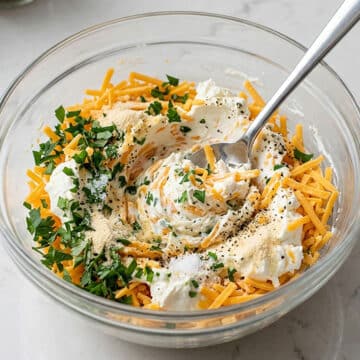 Cream cheese, cheddar cheese, parsley, garlic powder, onion powder, salt, pepper being mixed together in a glass bowl.