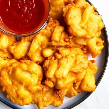 Easy One Pot Crispy Onion Fritters (Indian Pakoda) in White Dish With Bowl of Red Dipping Sauce.