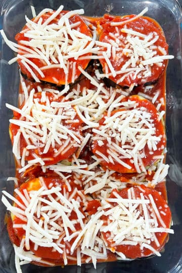 Lots of shredded cheese added to top of marinara covered eggplant slices in glass baking dish. 